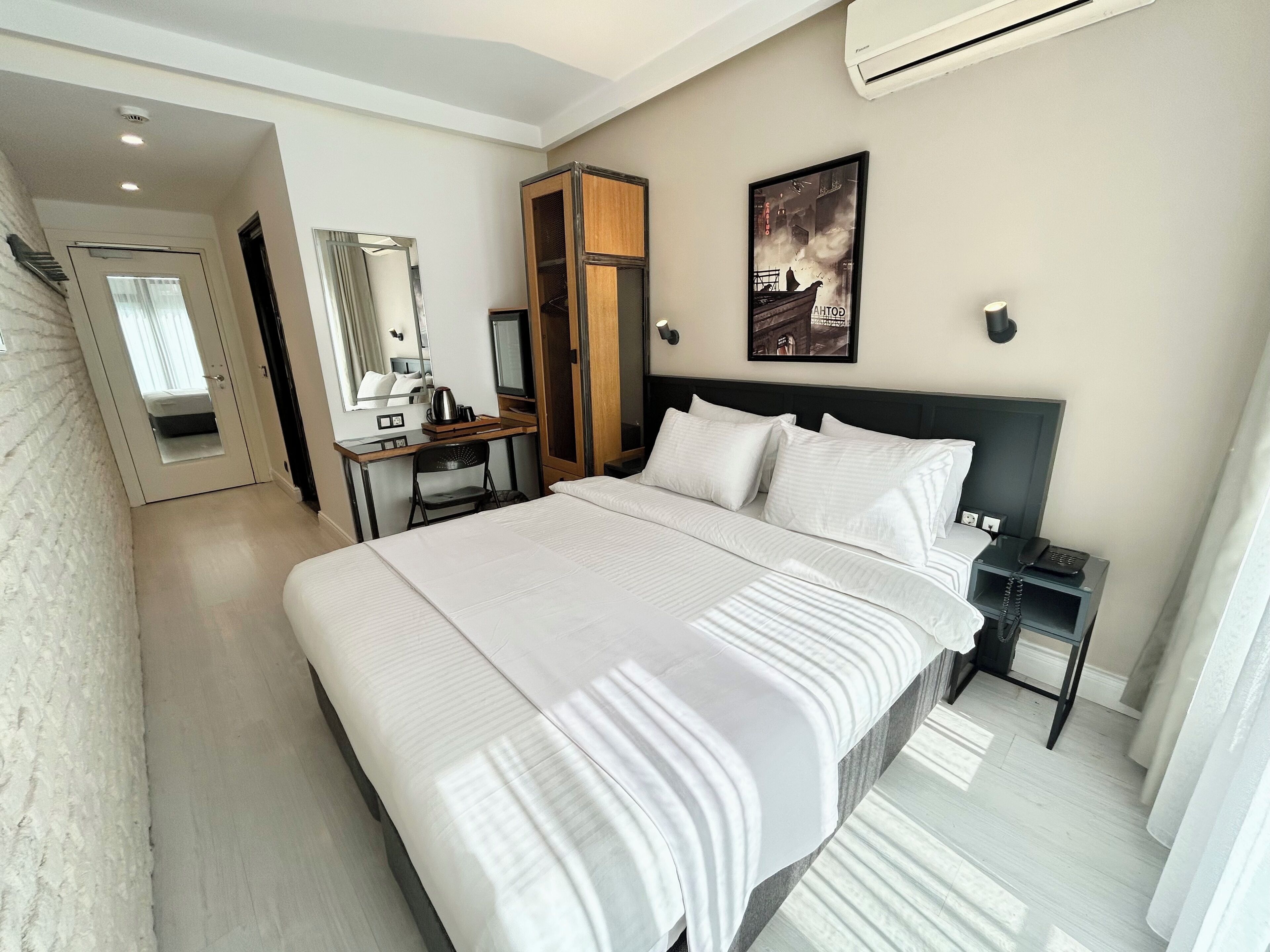 Une chambre d’hôtel moderne avec un grand lit, un bureau avec une chaise, une armoire, un miroir et un tableau encadré au mur.