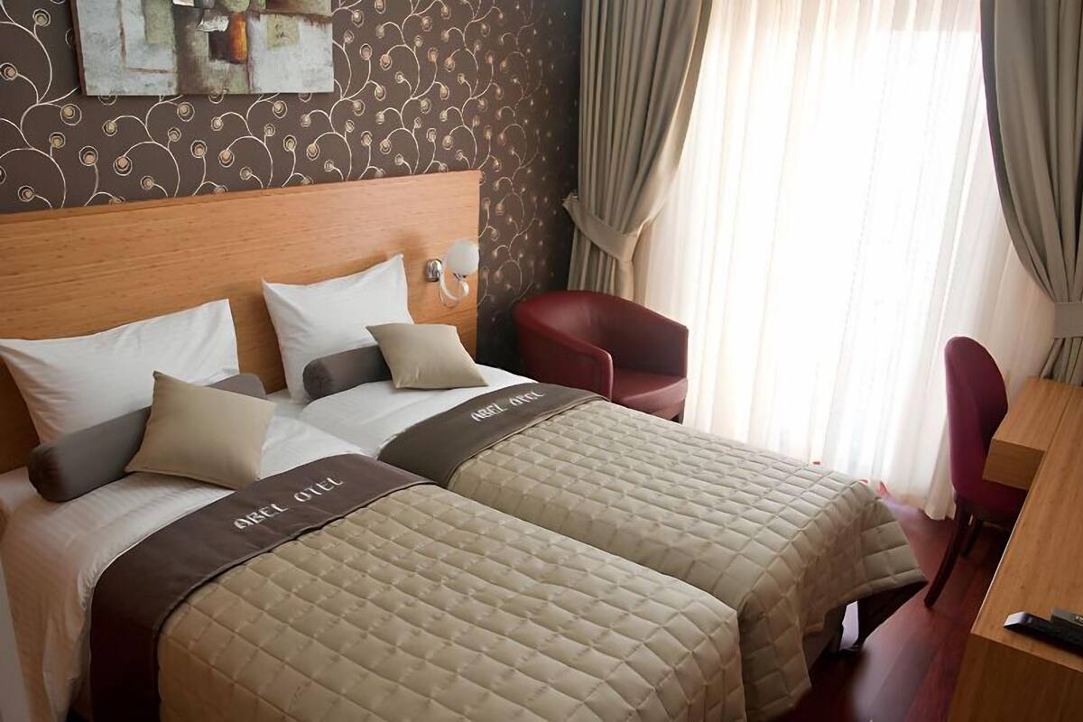 A hotel room with two beds, a wooden headboard, a red armchair, a desk, and a window with curtains.