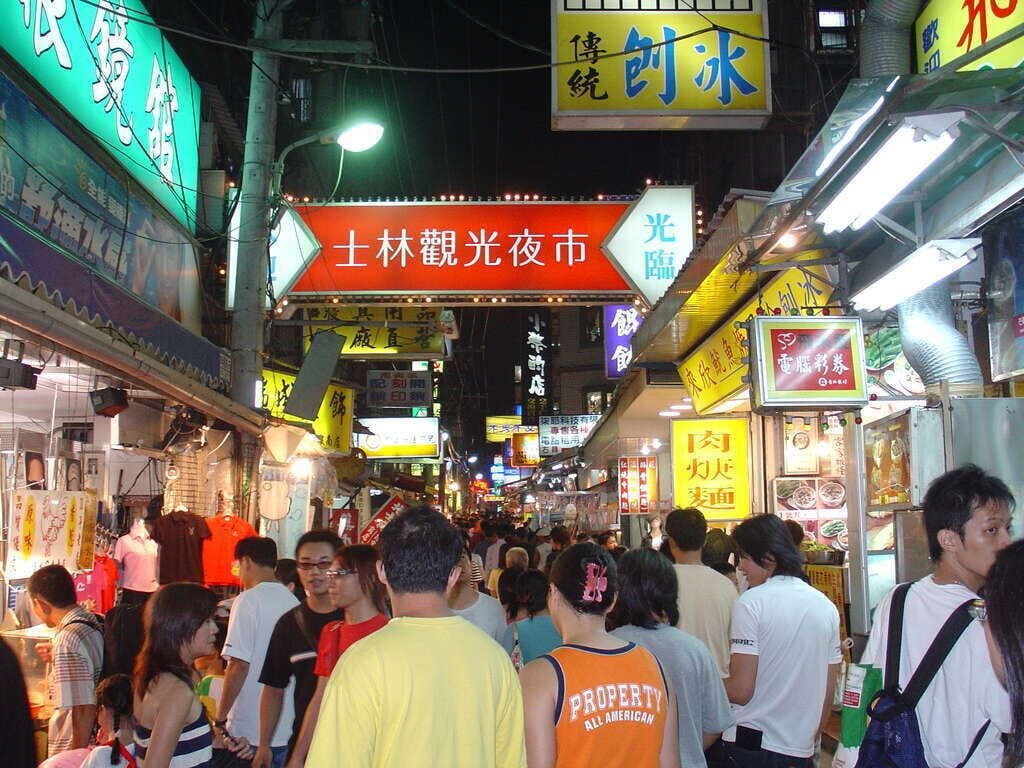 A crowded street with various shops and signs, including a prominent red sign with white text.