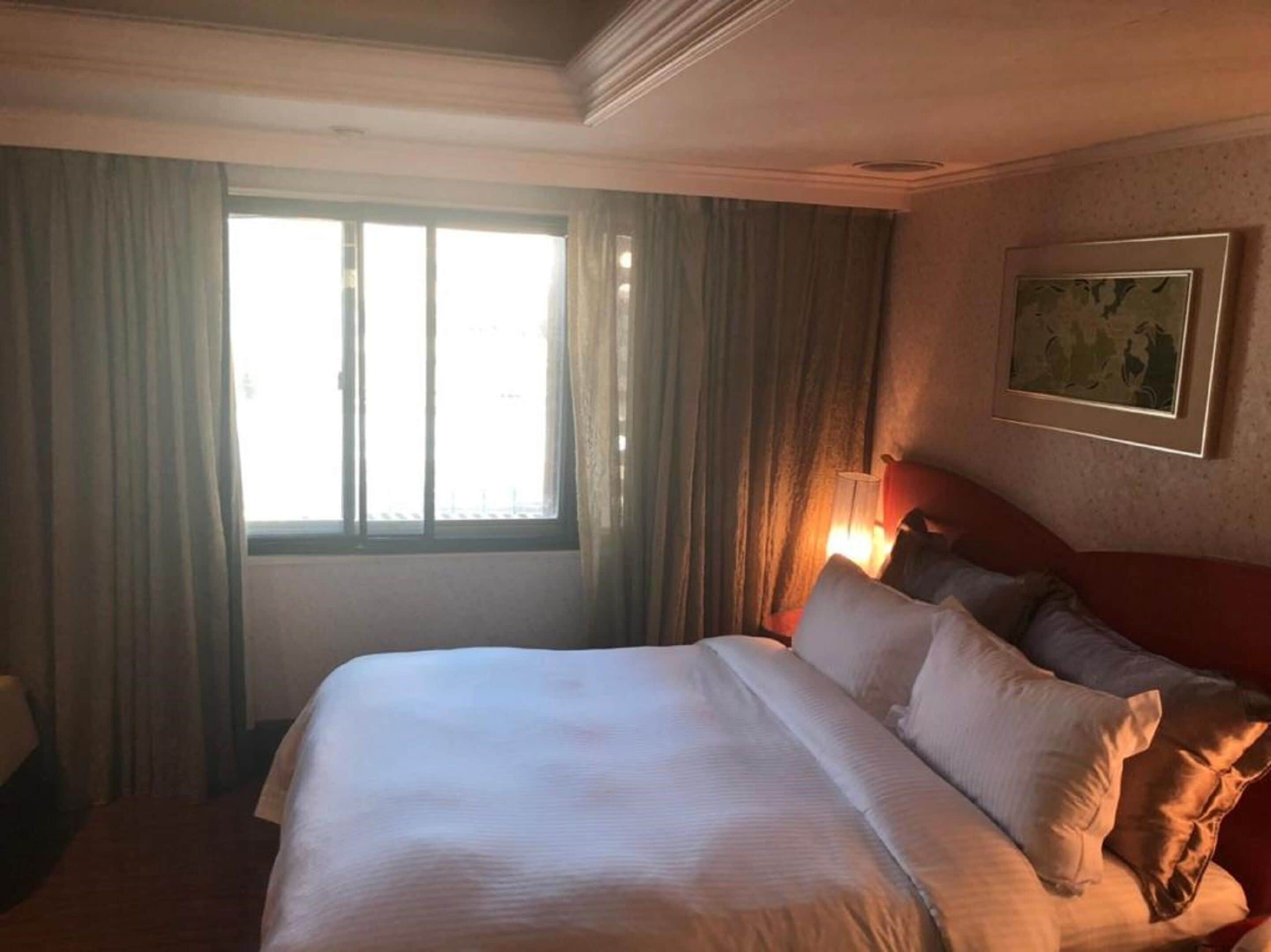 A hotel room with a large bed, a window with curtains, a wall-mounted painting, and a bedside lamp.