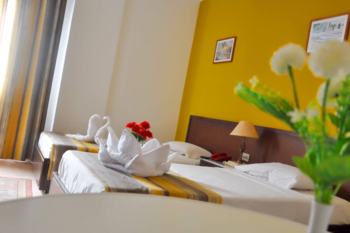 A hotel room with two beds, a wooden headboard, a bedside table with a lamp, a telephone, and a vase with flowers.