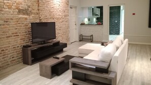 Un salon moderne avec un mur en briques, un téléviseur à écran plat, un canapé et une cuisine en arrière-plan.