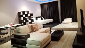 Un salon moderne avec un canapé, une table basse et un téléviseur. On y trouve un lit avec une tête de lit noire et une grande fenêtre avec des rideaux.