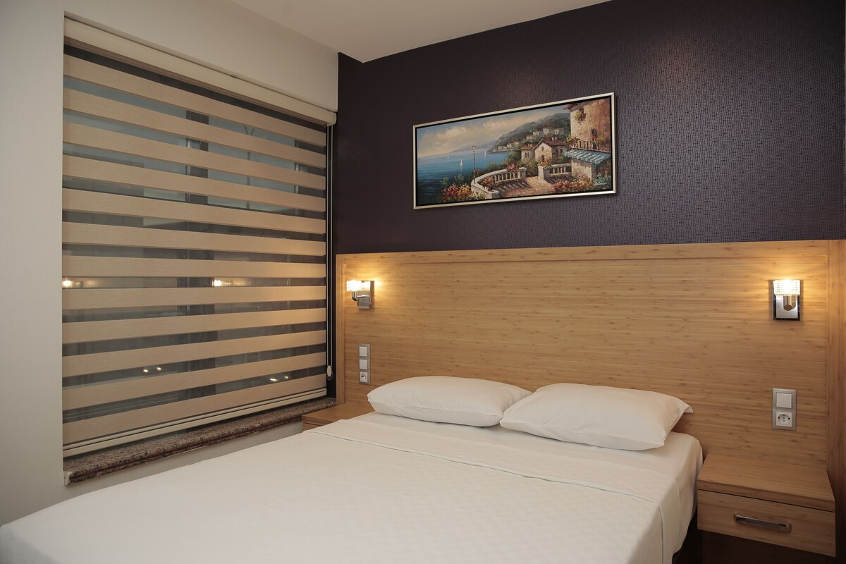 Habitación de hotel con una cama, cabecera de madera, mesita de noche, un cuadro colgado en la pared y una ventana con persianas.