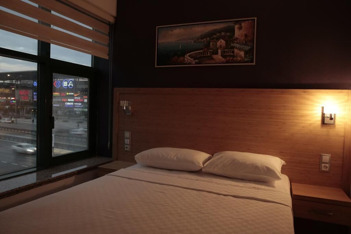 Habitación de hotel con una cama grande, una mesita de noche, una lámpara de pared y una ventana con vista a la ciudad.