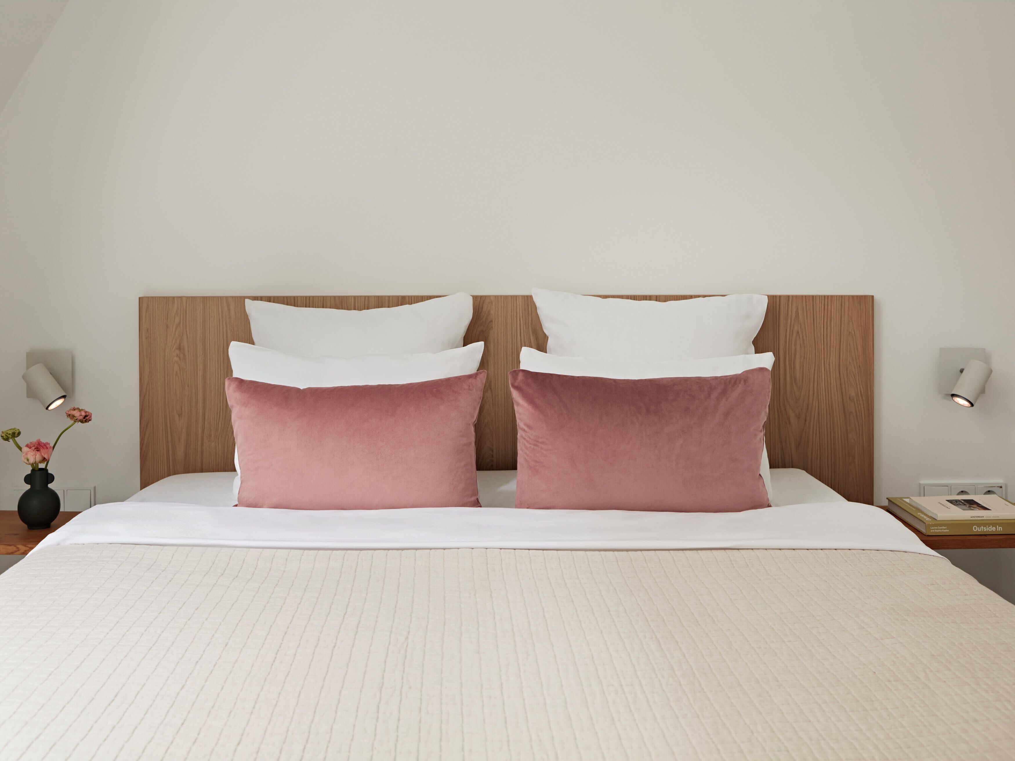 A neatly made bed with two pillows, a bedside table with a lamp, and a wall-mounted light fixture.