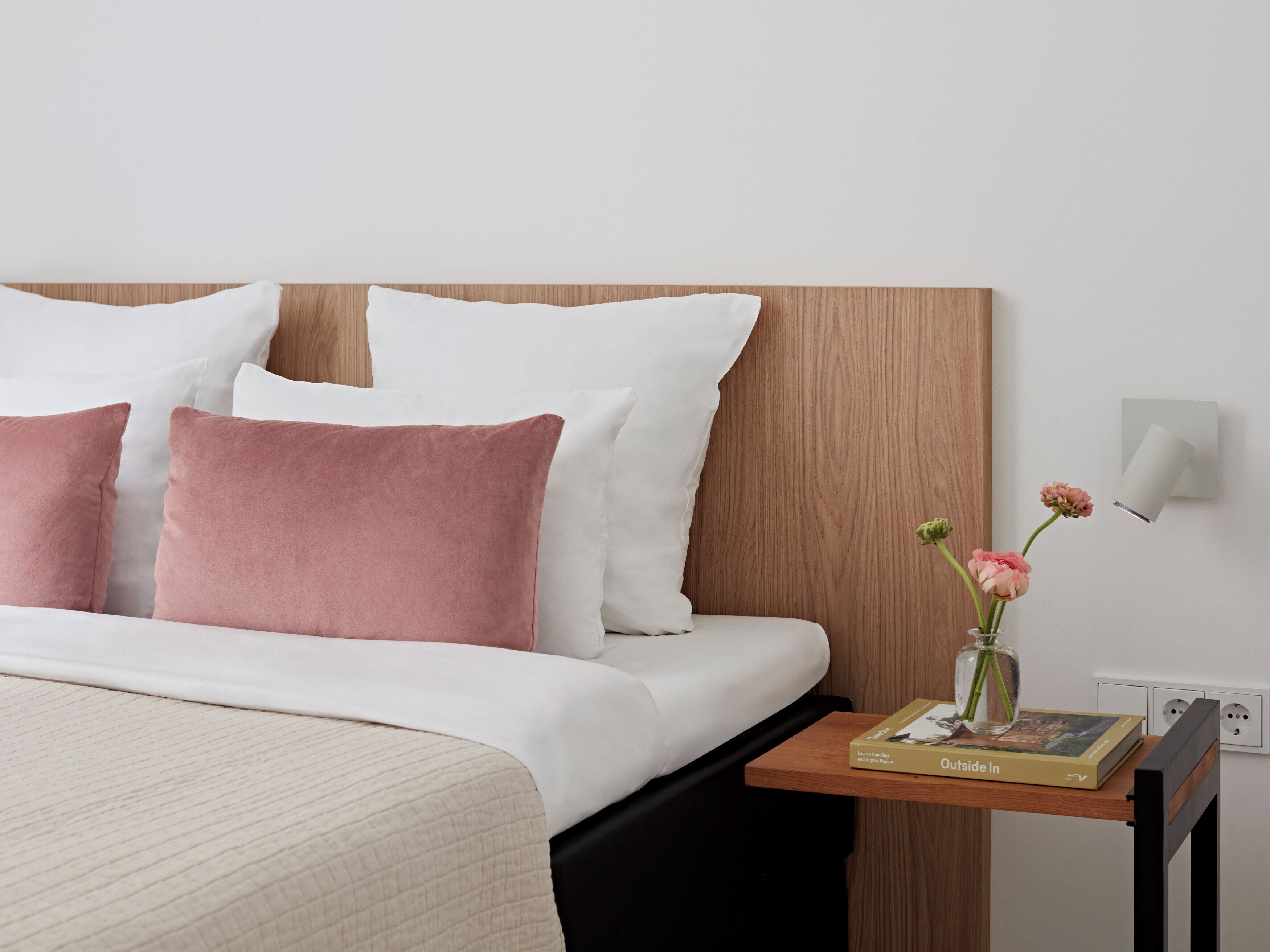 Una cama bien hecha con almohadas blancas y rosas, una mesita de noche con un libro y un jarrón con flores.