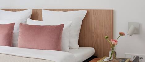 Una cama bien hecha con almohadas blancas y rosas, una mesita de noche con un libro y un jarrón con flores.