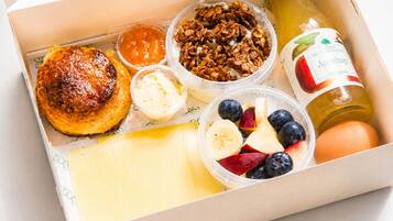 A breakfast box containing a croissant, granola, yogurt, fruit, cheese, and a bottle of apple cider vinegar.