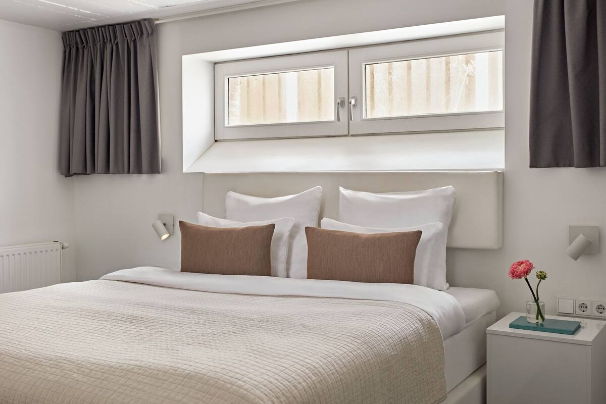 A neatly arranged bedroom with a bed, pillows, a bedside table, a nightstand, a window, and a wall-mounted light fixture.