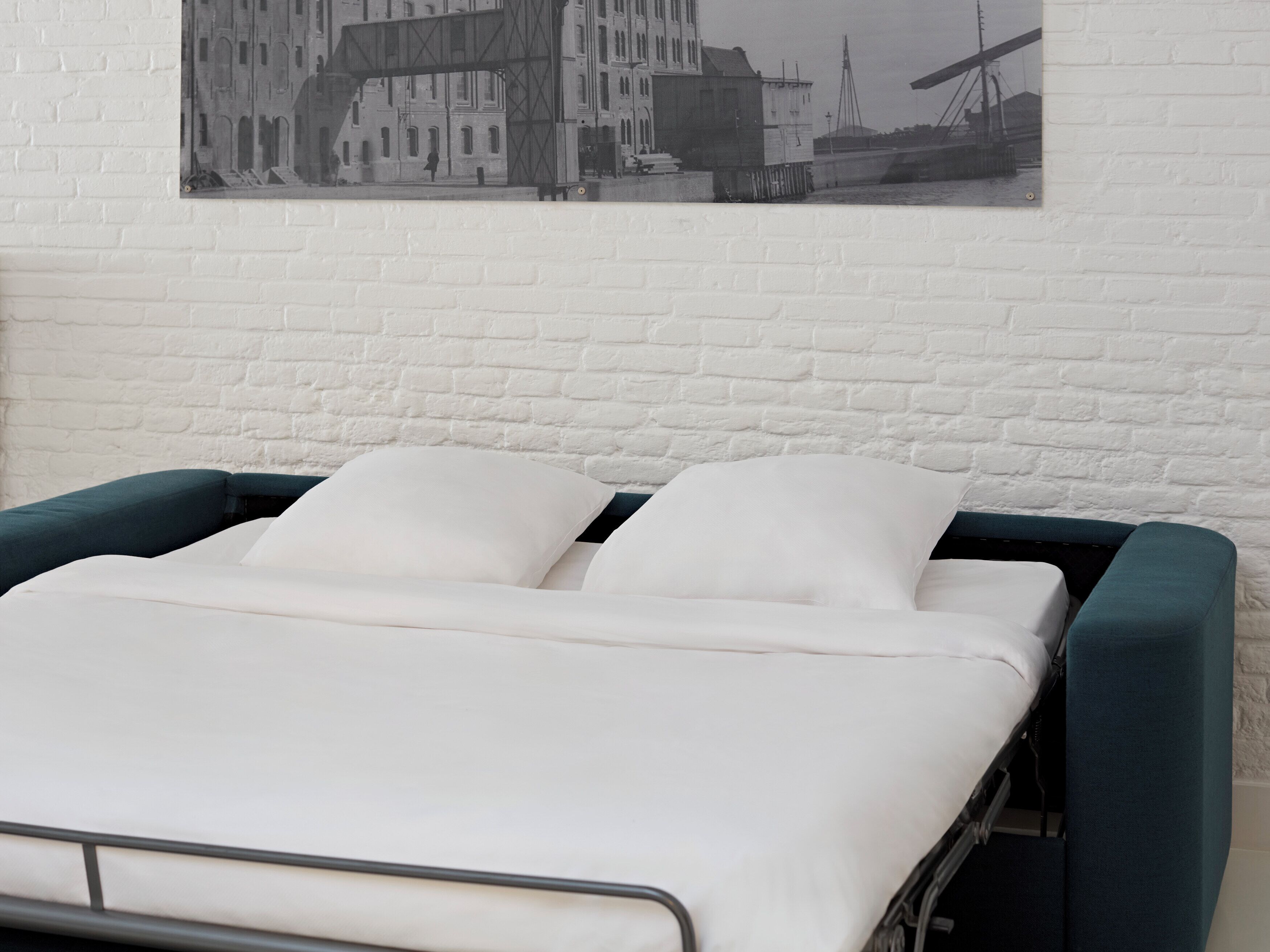 Ein Bett mit weißer Bettwäsche, eine weiße Ziegelwand und ein Schwarz-Weiß-Foto eines Gebäudes.