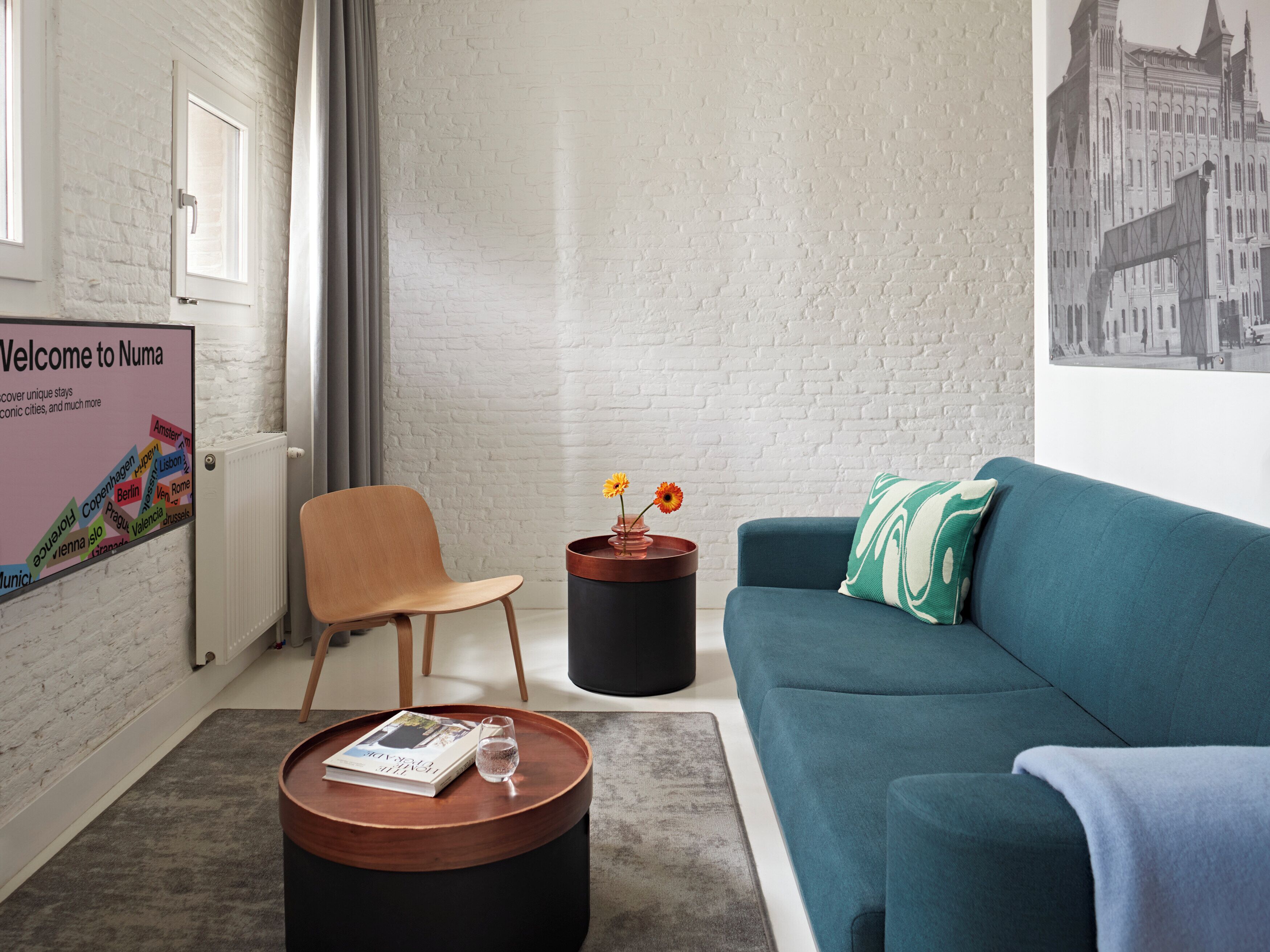 Un salon moderne avec un canapé bleu, une chaise en bois, une table basse ronde sur laquelle se trouvent un livre et un verre, et une pancarte « Bienvenue chez Numa » accrochée au mur.