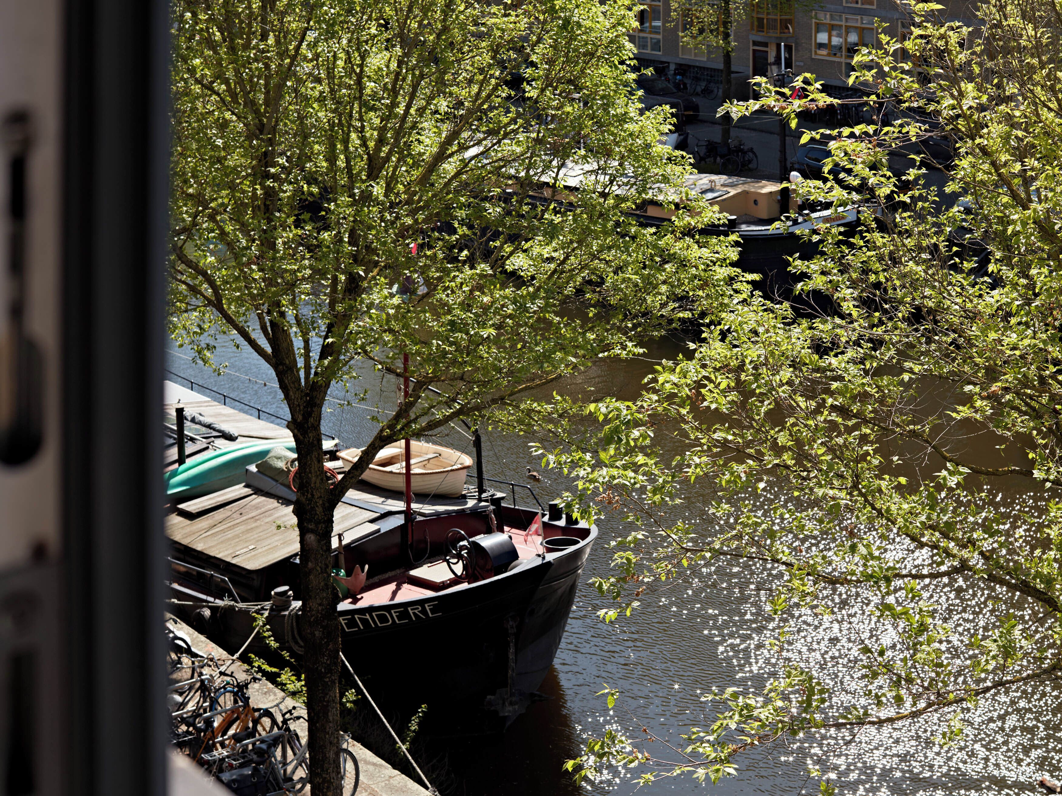 A boat docked at a pier with bicycles parked nearby, a tree in the foreground, and a building in the background.