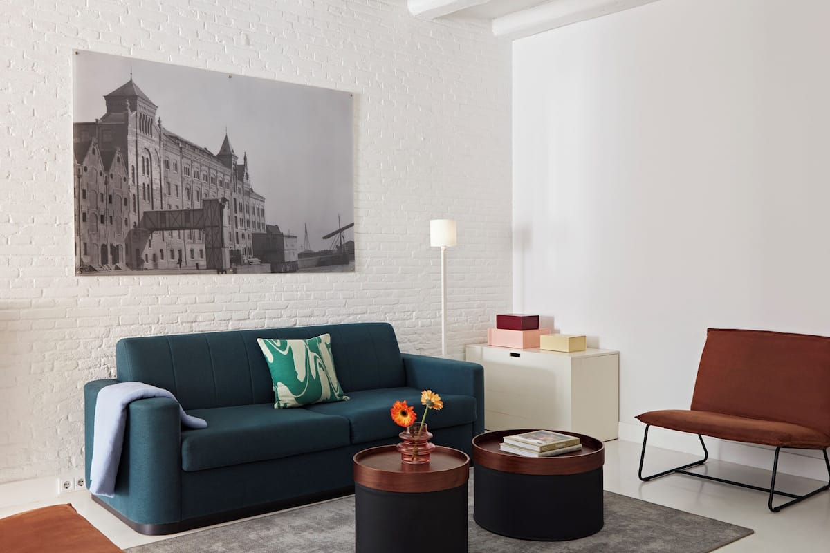 A modern living room with a teal sofa, two round coffee tables, a brown leather armchair, a white side table with boxes, and a cityscape painting on the wall.