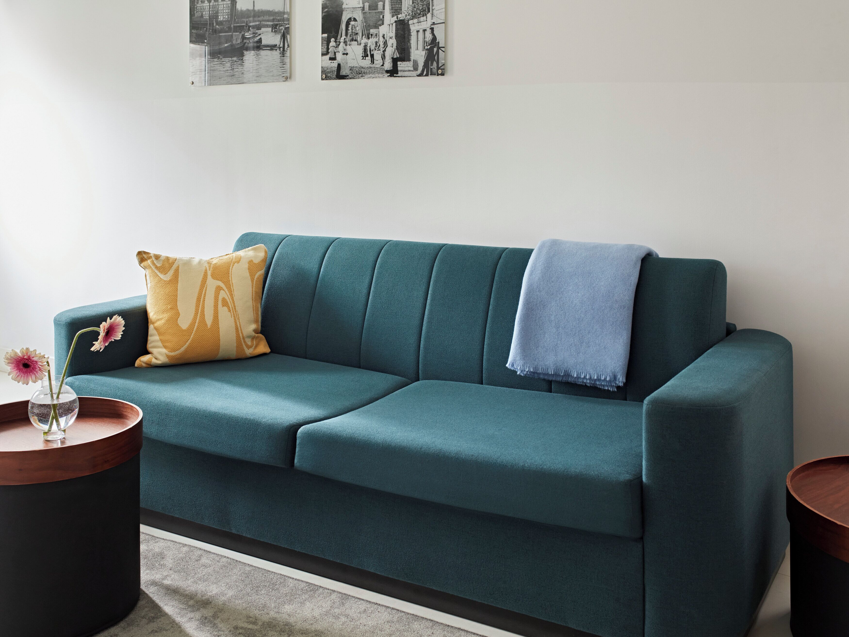 A teal sofa with a yellow pillow, a blue throw, and a vase with flowers on a wooden side table.