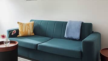 Un sofá color turquesa con un cojín amarillo, una manta azul y un jarrón con flores sobre una mesita de madera.