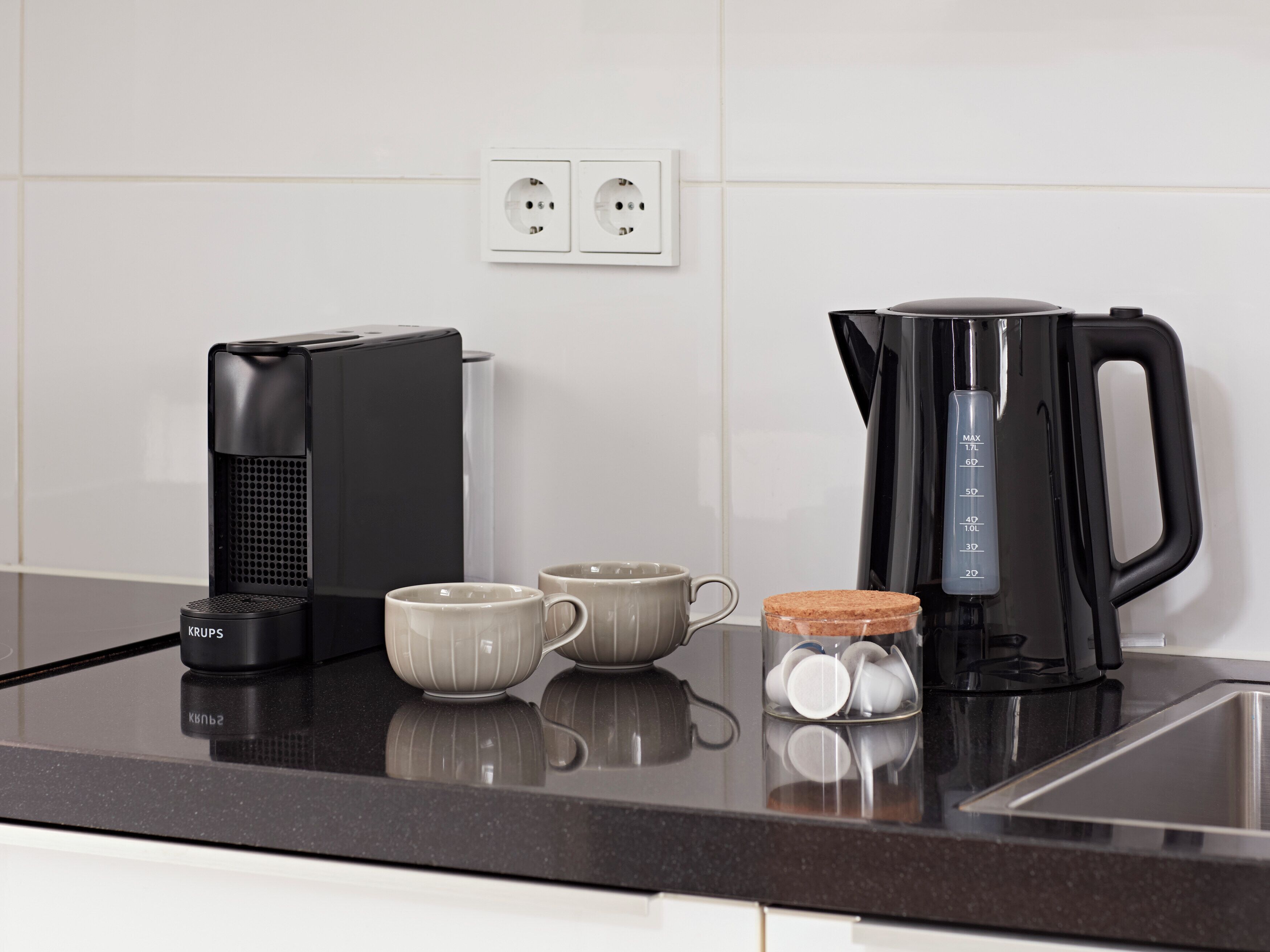 Una encimera de cocina con una cafetera, dos tazas, un hervidor y un tarro de azúcar.