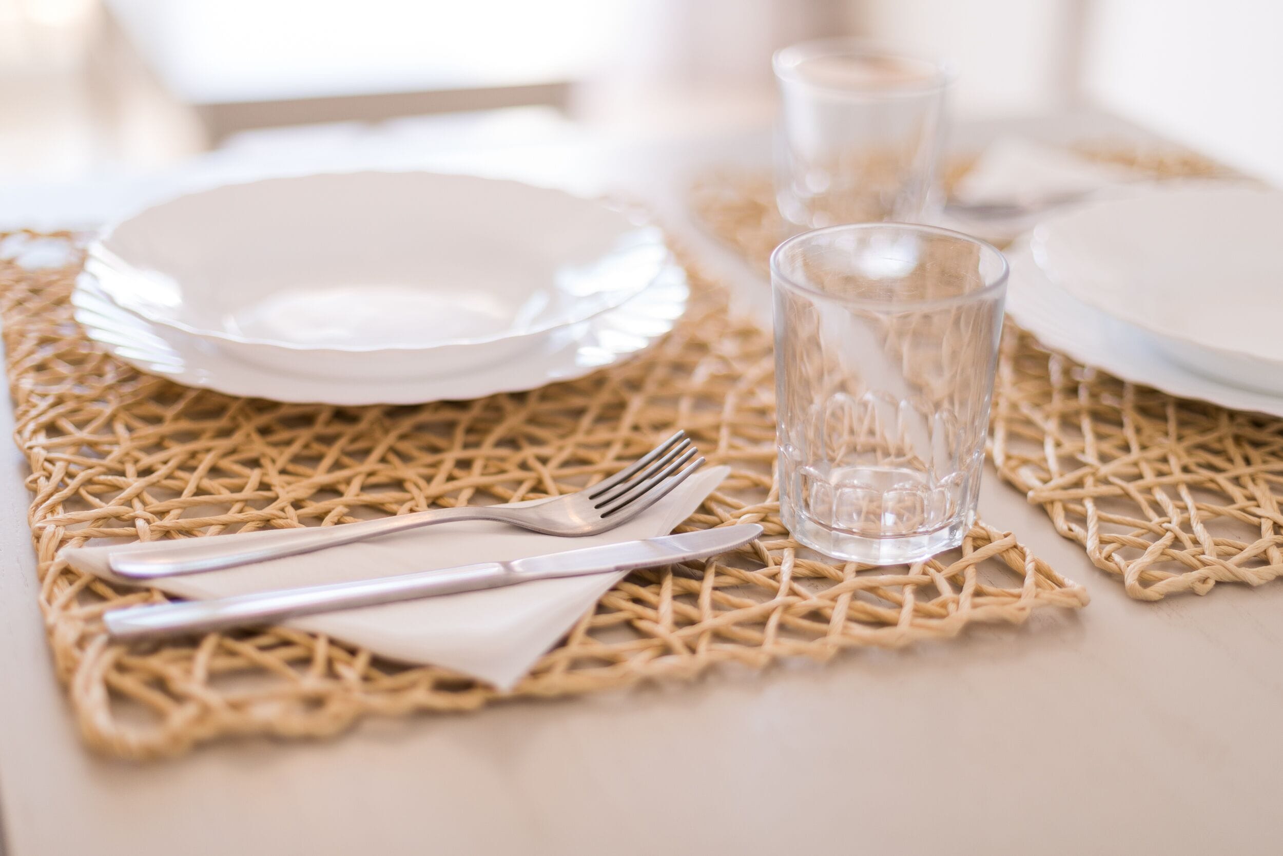 Une table dressée avec des assiettes blanches, un verre transparent et des couverts en argent, le tout posé sur un set de table en fibres tressées.