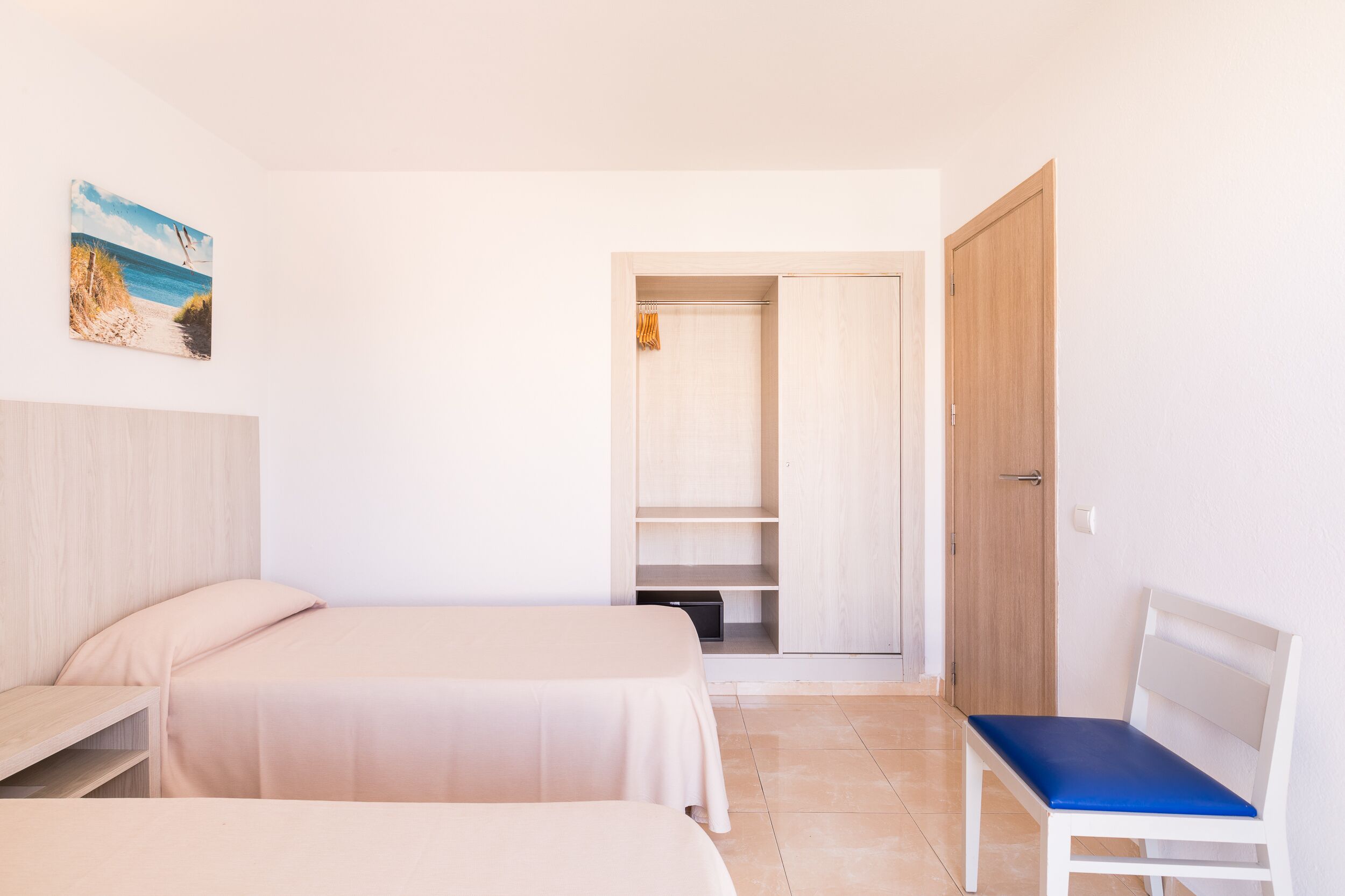 Un dormitorio con dos camas, un banco, un armario y un cuadro de una playa.