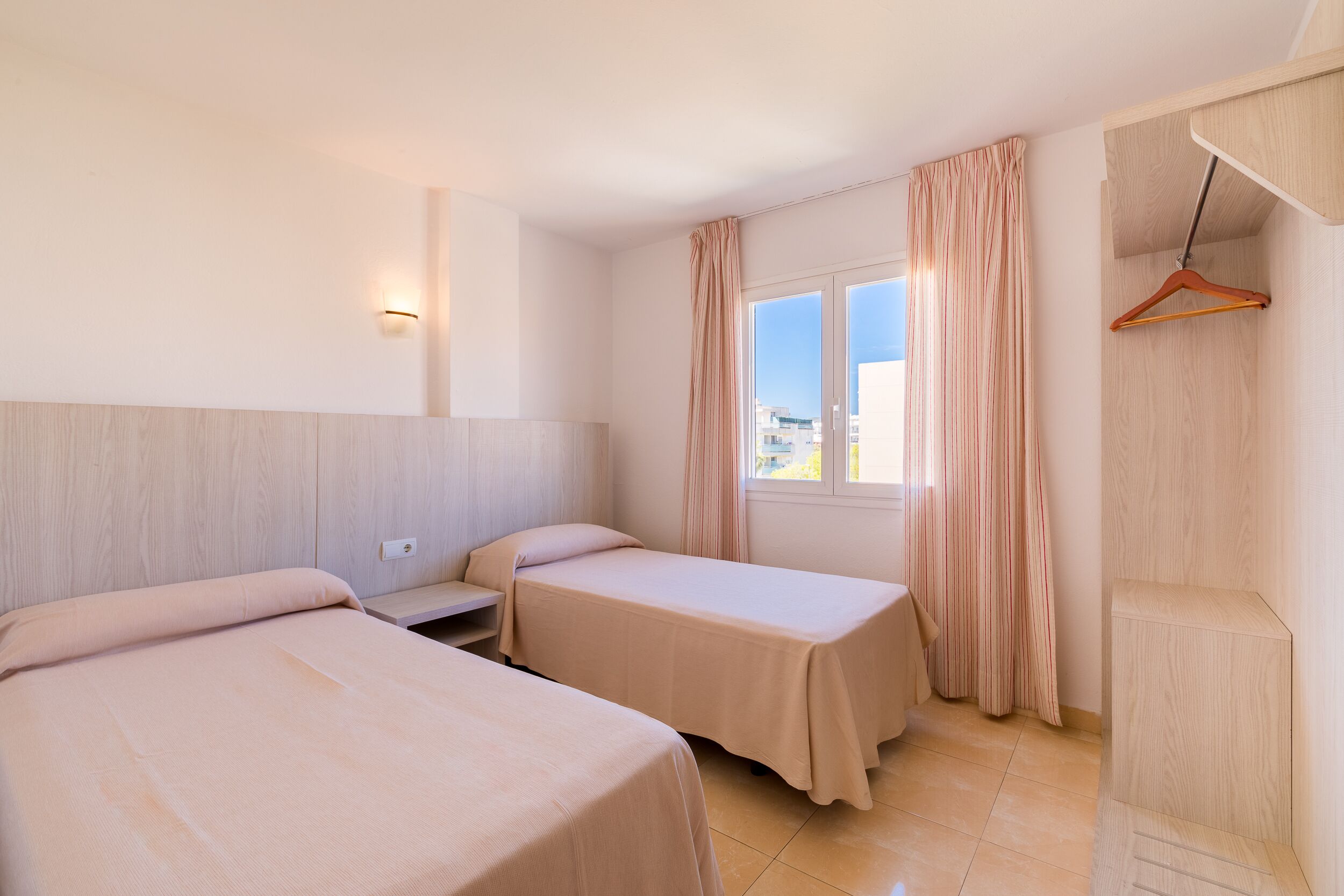 A hotel room with two beds, a wooden headboard, a window with curtains, and a wooden wardrobe.