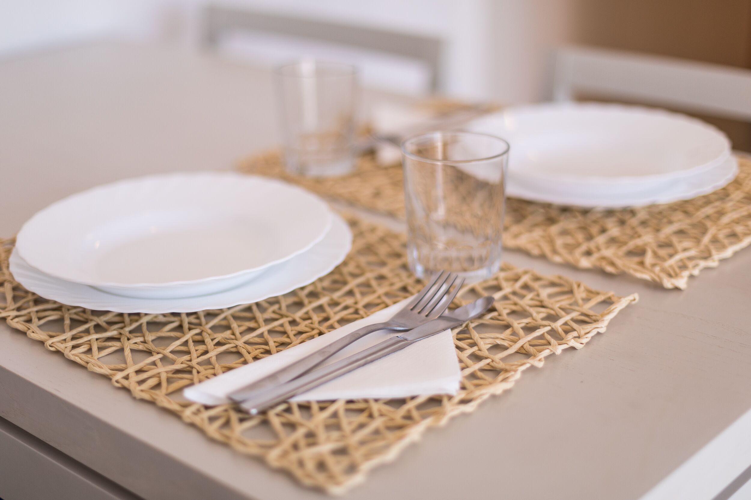 Una mesa de comedor con platos blancos, cubiertos y un vaso sobre un camino de mesa tejido.