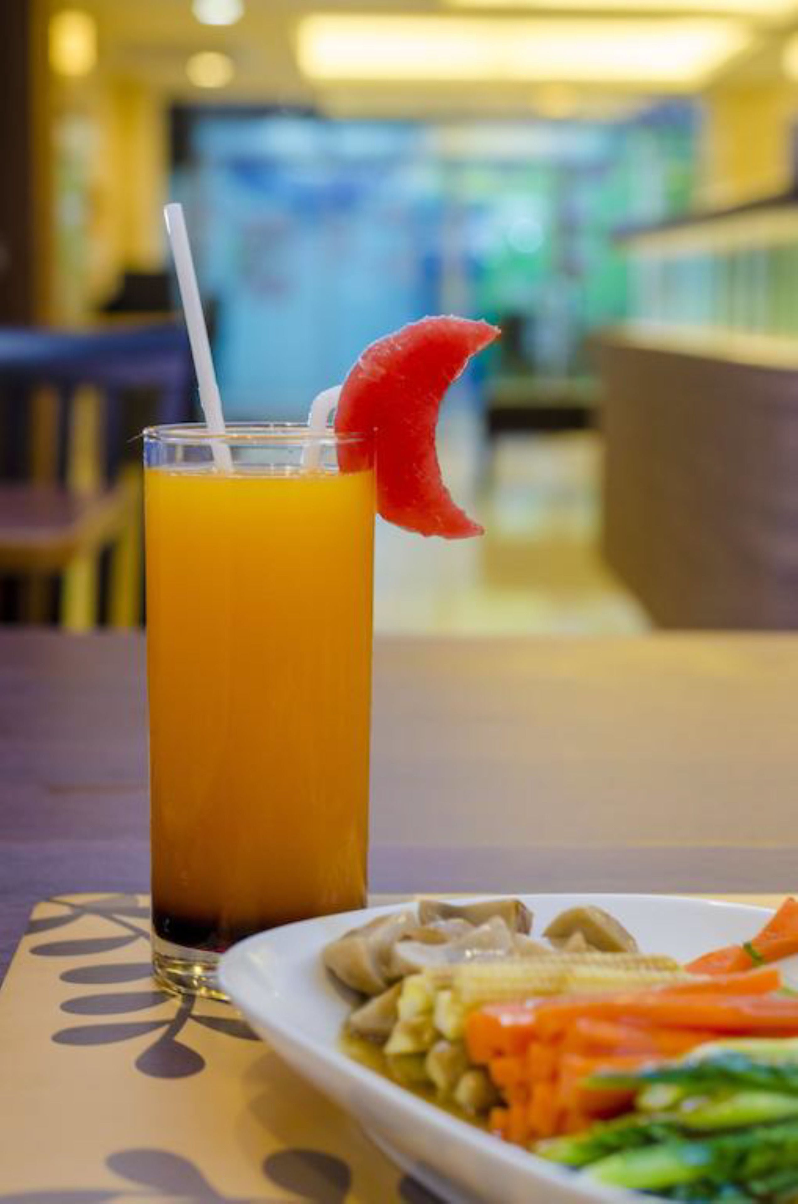 A glass of orange juice with a straw and a slice of red fruit on the rim, placed on a table with a plate of vegetables.