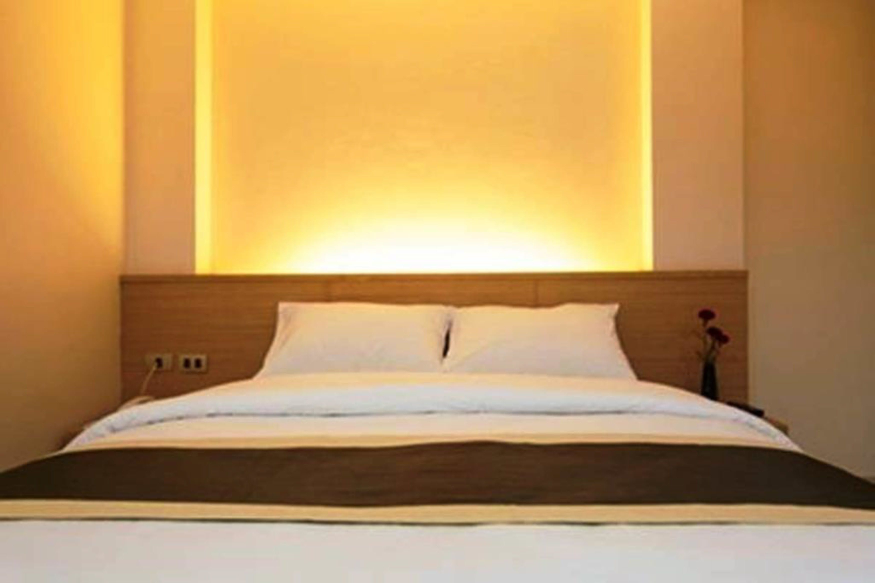 A neatly made bed with a wooden headboard, a bedside table with a vase of flowers, and a wall-mounted light fixture.