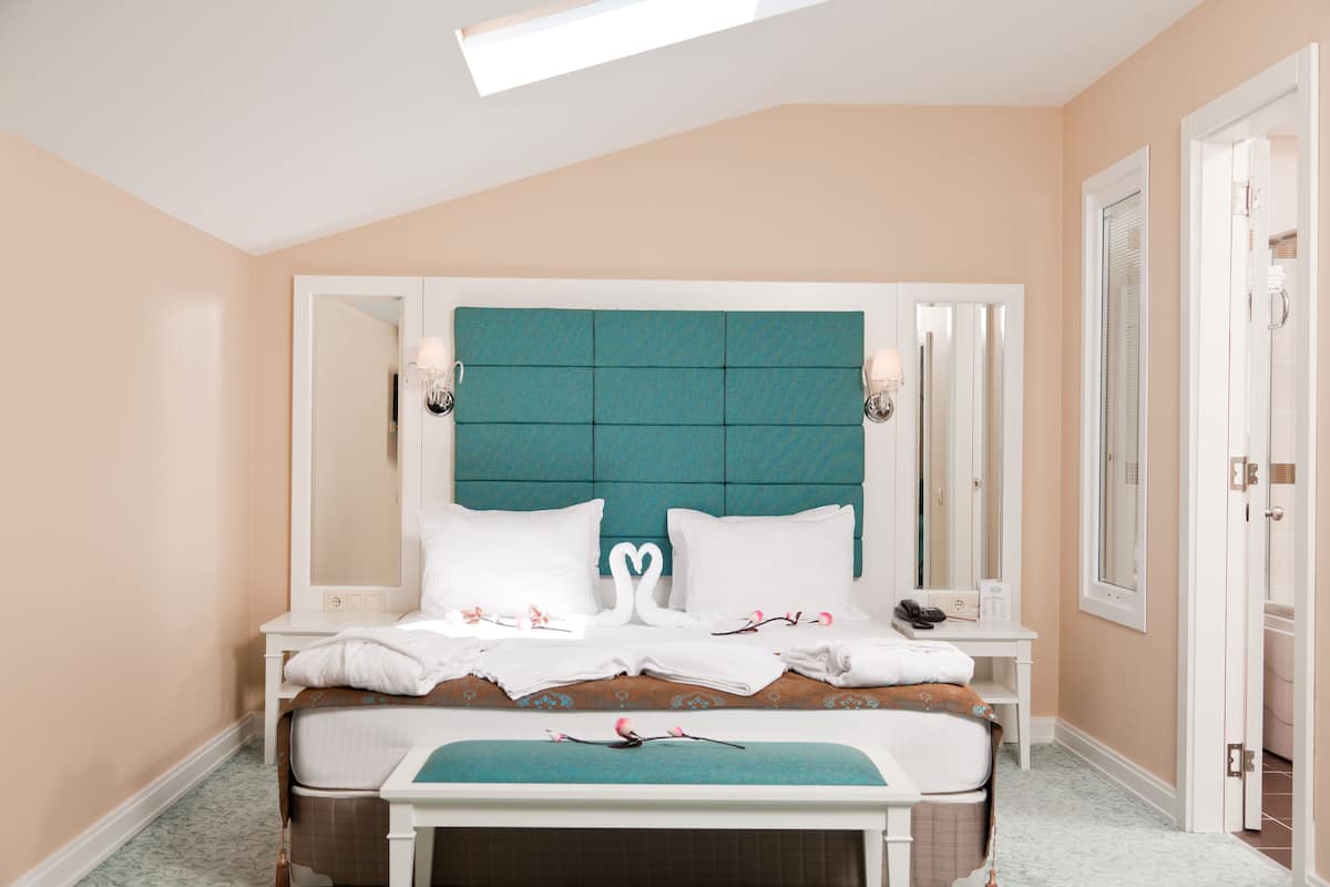 A neatly arranged hotel room with a teal headboard, white bedding, and a small desk with a phone and a lamp.