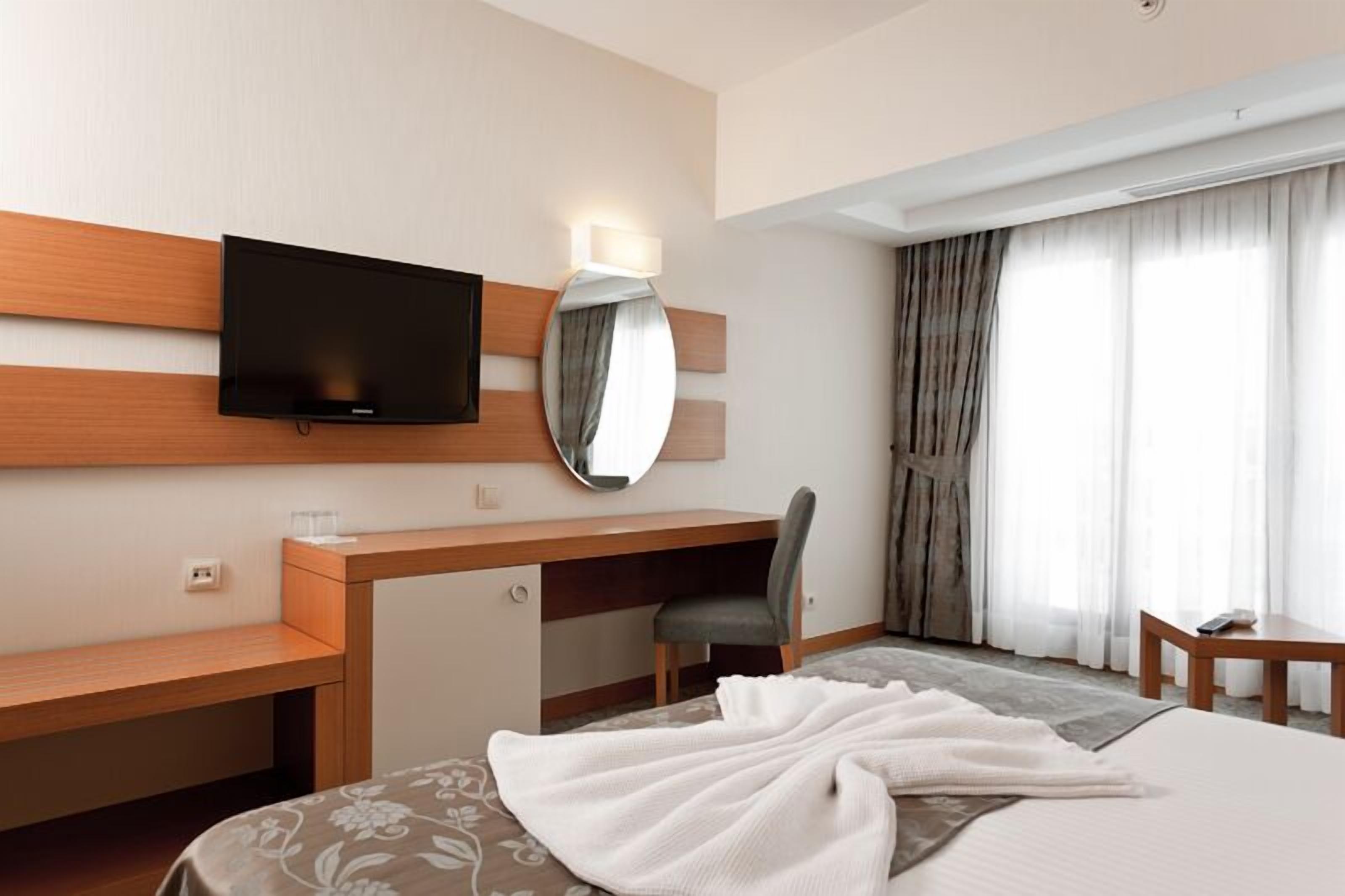 A hotel room with a bed, a desk, a chair, a TV, a mirror, and a window with curtains.