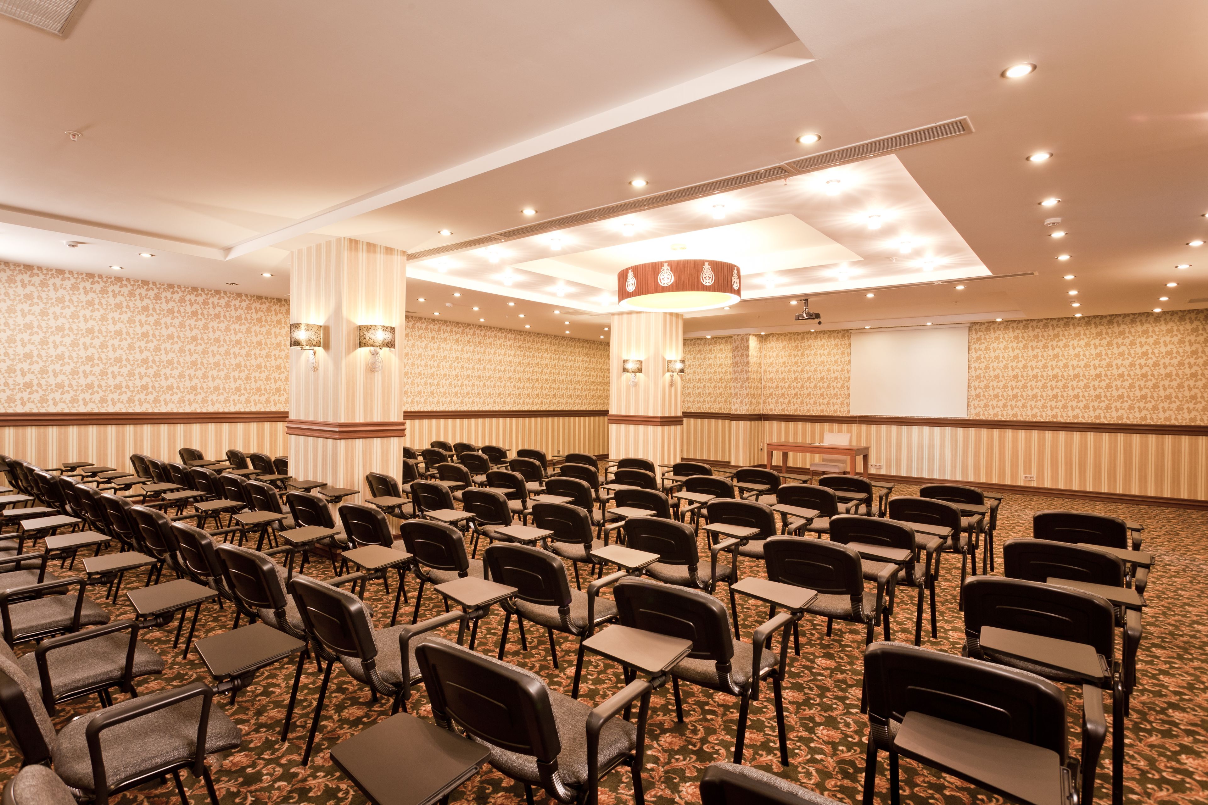 Un salón de conferencias con filas de sillas y mesas, una pantalla de proyector y un pilar grande con luminarias.