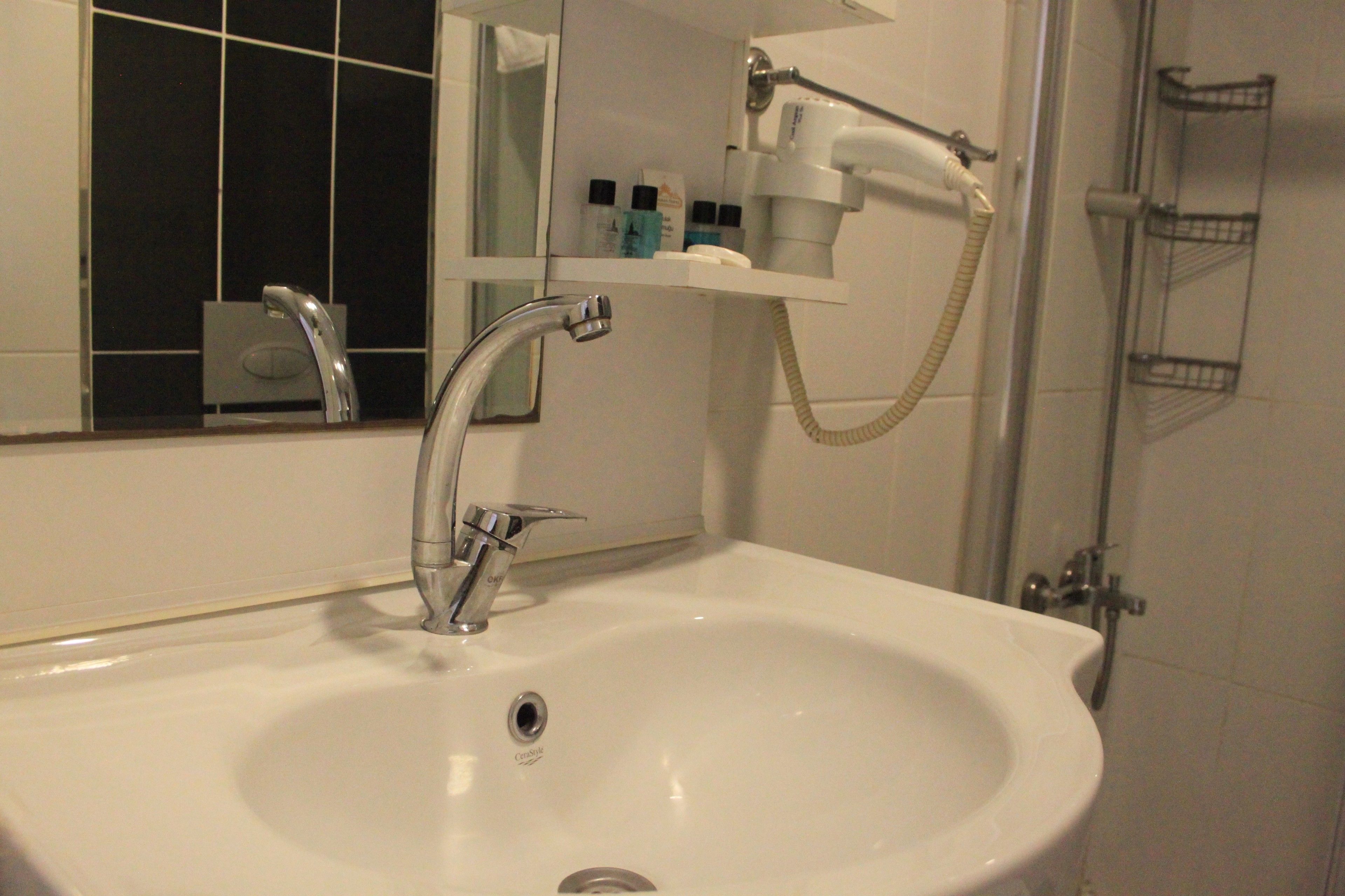 Ein Badezimmer mit einem weißen Waschbecken, einem Chromarmaturen-Set und einem Spiegel darüber. Ein Ablagebereich mit Pflegeprodukten und ein Duschbereich sind ebenfalls zu sehen.