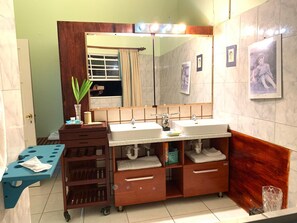A bathroom with a double sink, wooden vanity, a mirror, and a small table with a plant. - RNM The Clubhouse Grenada (St. John)