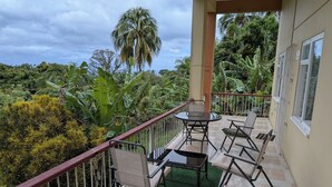 A balcony with a table and chairs, surrounded by lush greenery and a palm tree. - RNM The Clubhouse Grenada (St. John)