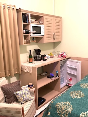 A compact room with a kitchenette, including a microwave, coffee maker, and a small refrigerator. There is a bed with a green patterned bedspread, a chair with a teddy bear, and a small table with a bottle and glasses. - RNM The Clubhouse Grenada (St. John)