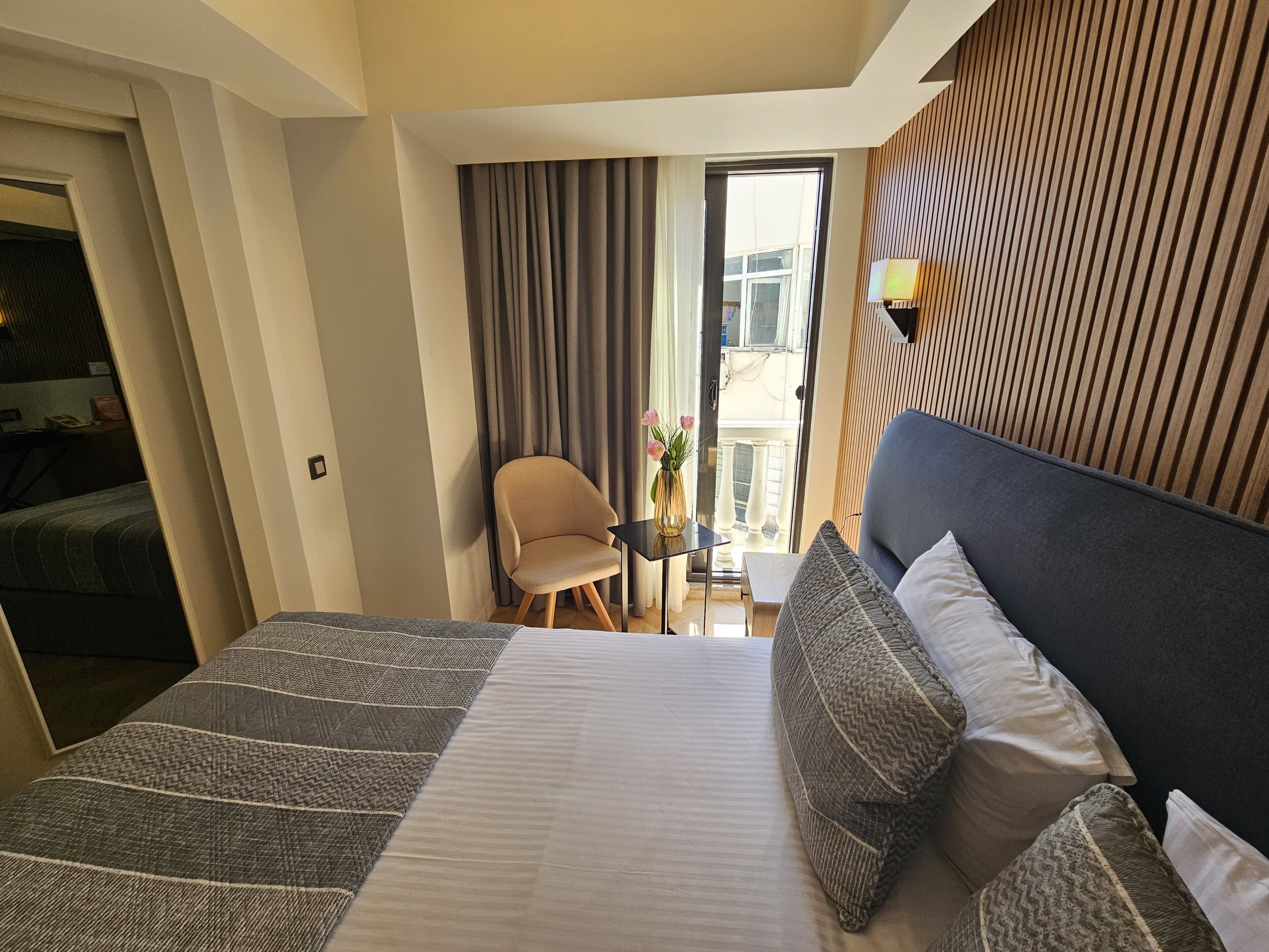 A hotel room with a bed, a chair, a small table with a vase of flowers, and a balcony with curtains.