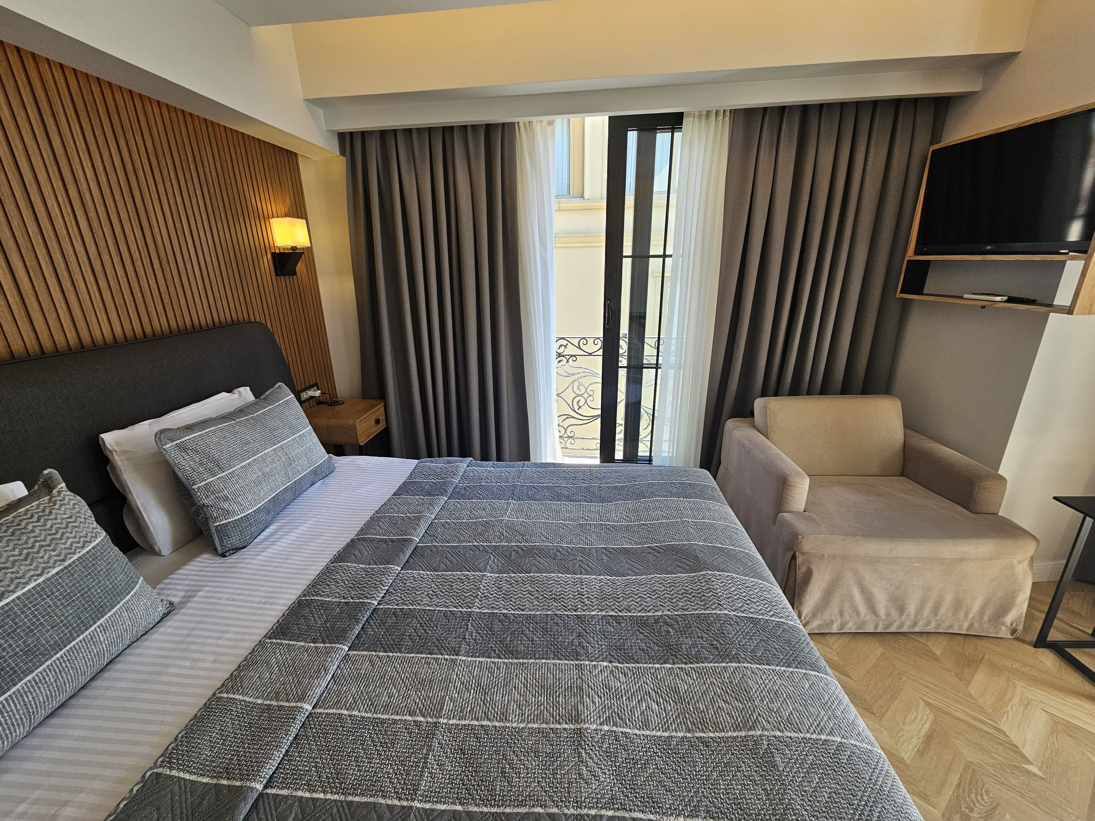 Una habitación de hotel con una cama grande, un sofá, un televisor montado en la pared y un balcón con cortinas.