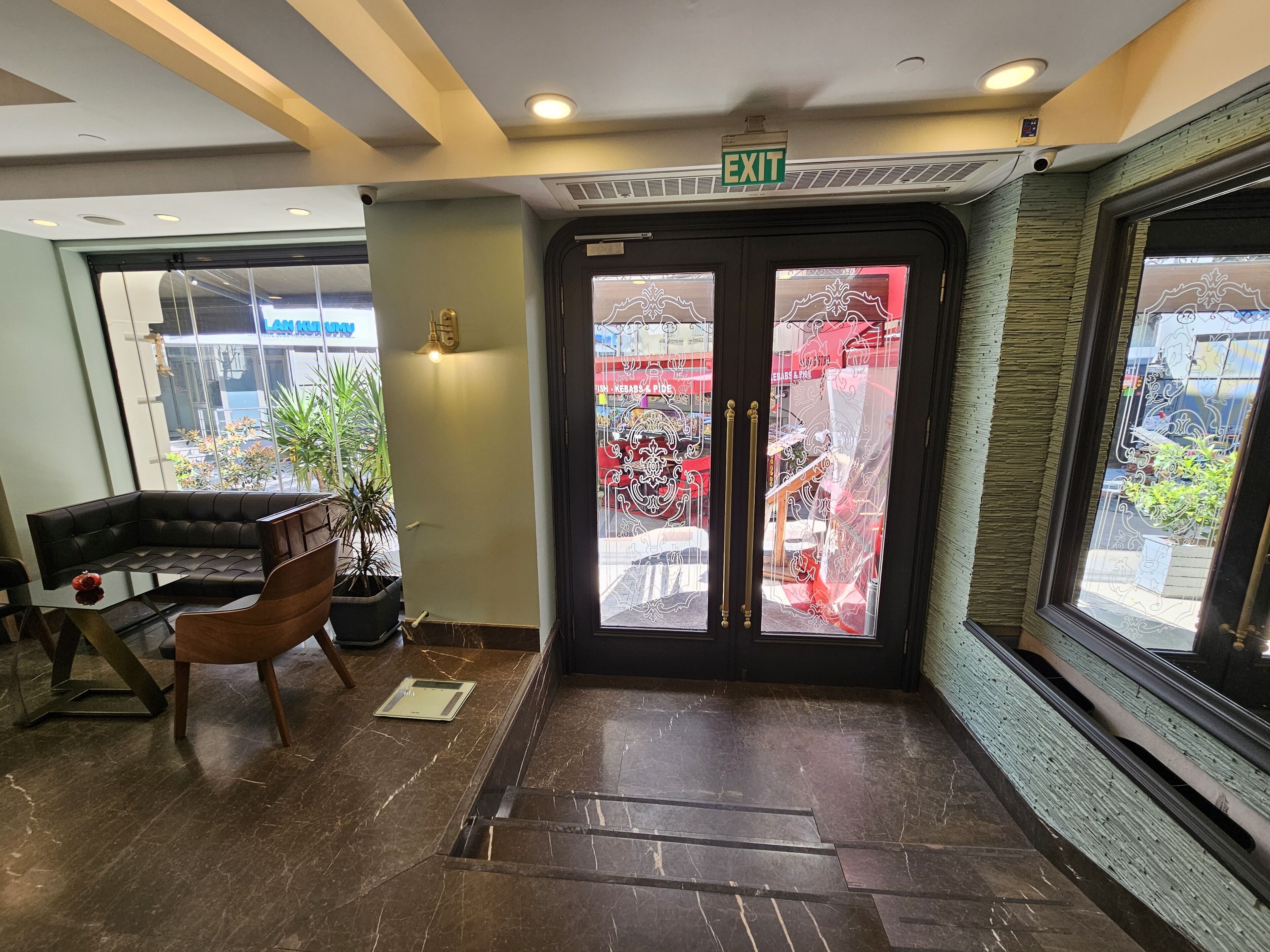 A hotel lobby with a reception desk, a sofa, a potted plant, and a glass door leading outside.