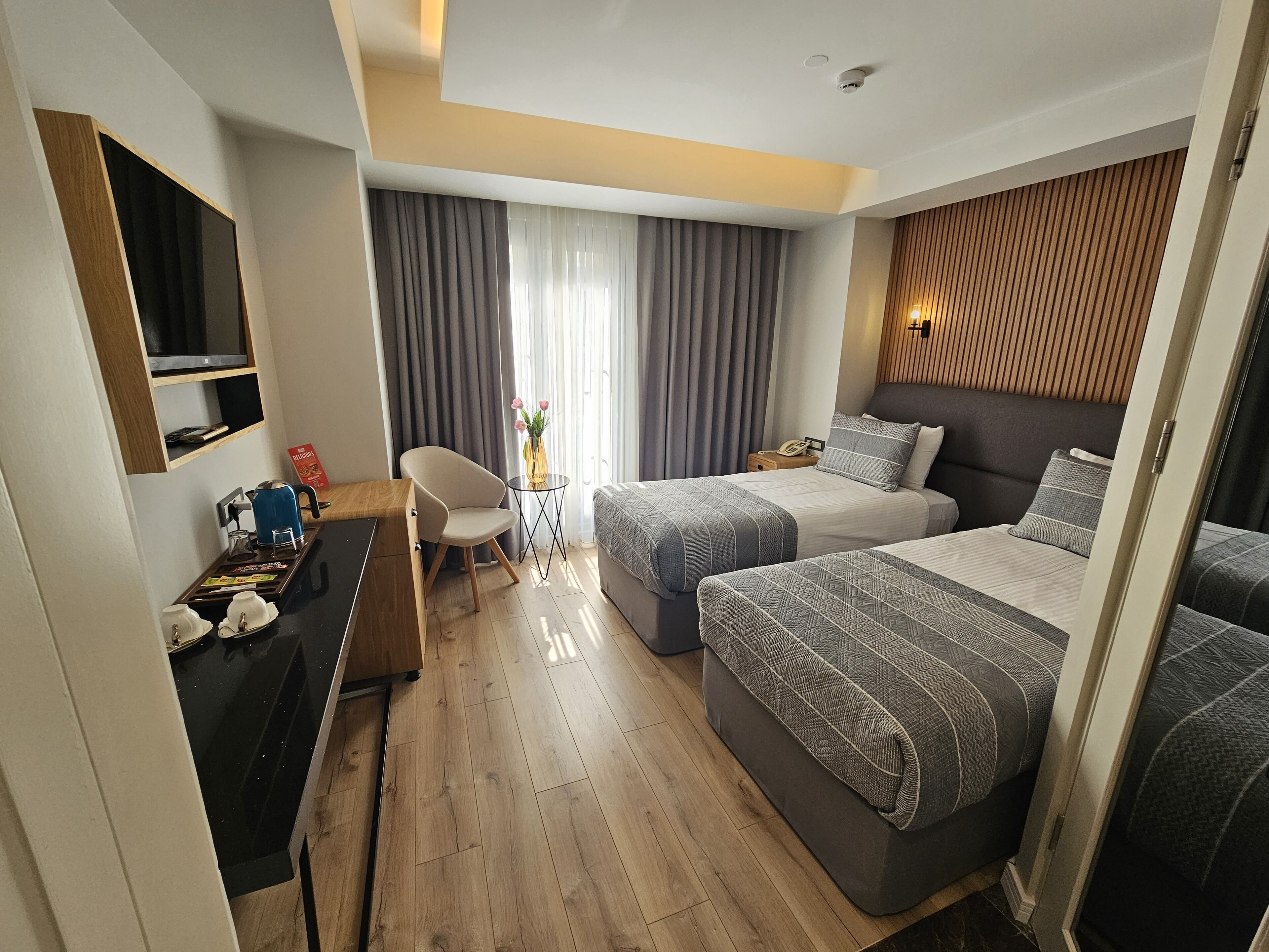 A modern hotel room with two beds, a desk with a chair, a small table with a vase, and a large window with curtains.