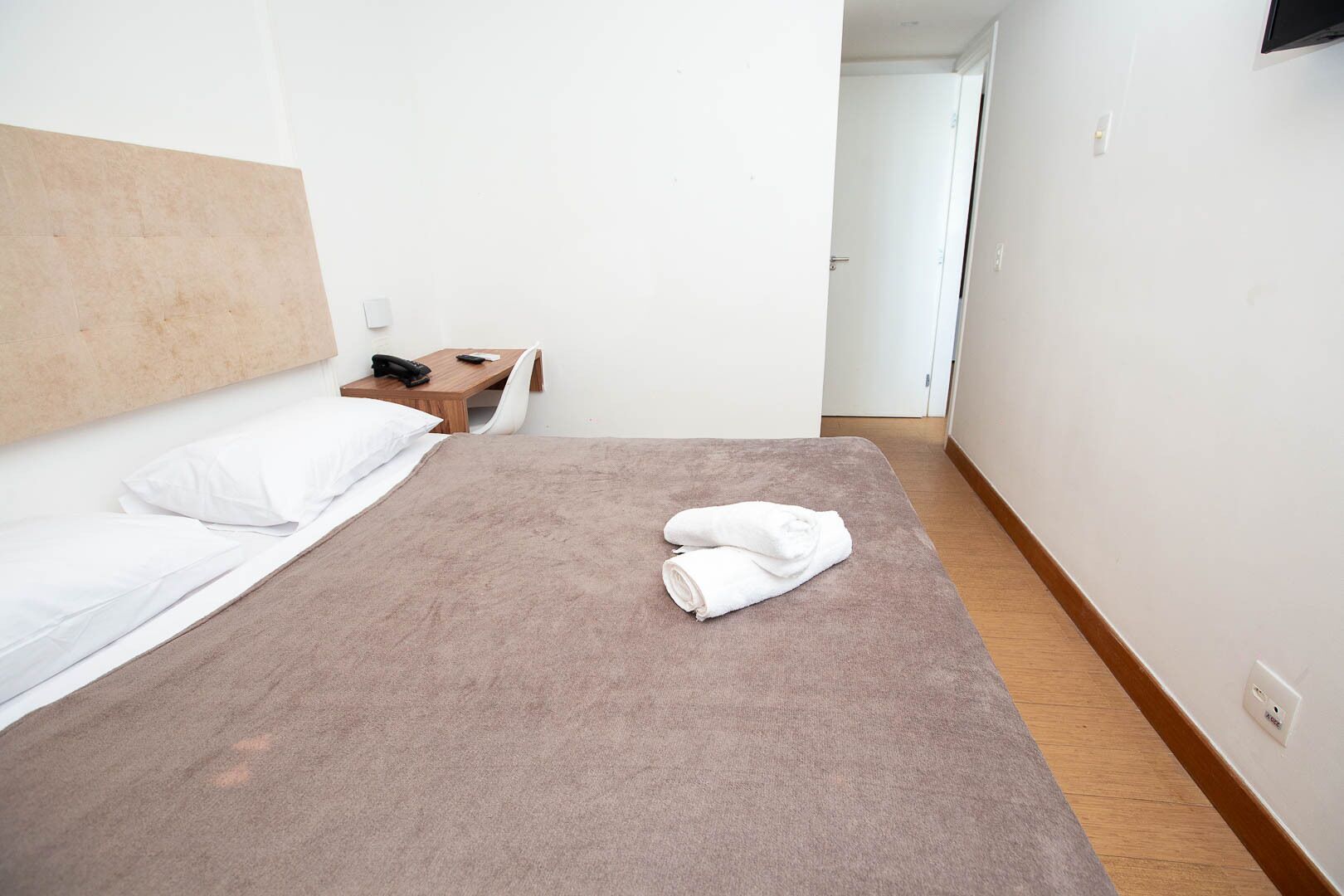 A hotel room with a bed, a bedside table with a phone and remote, and a door in the background.