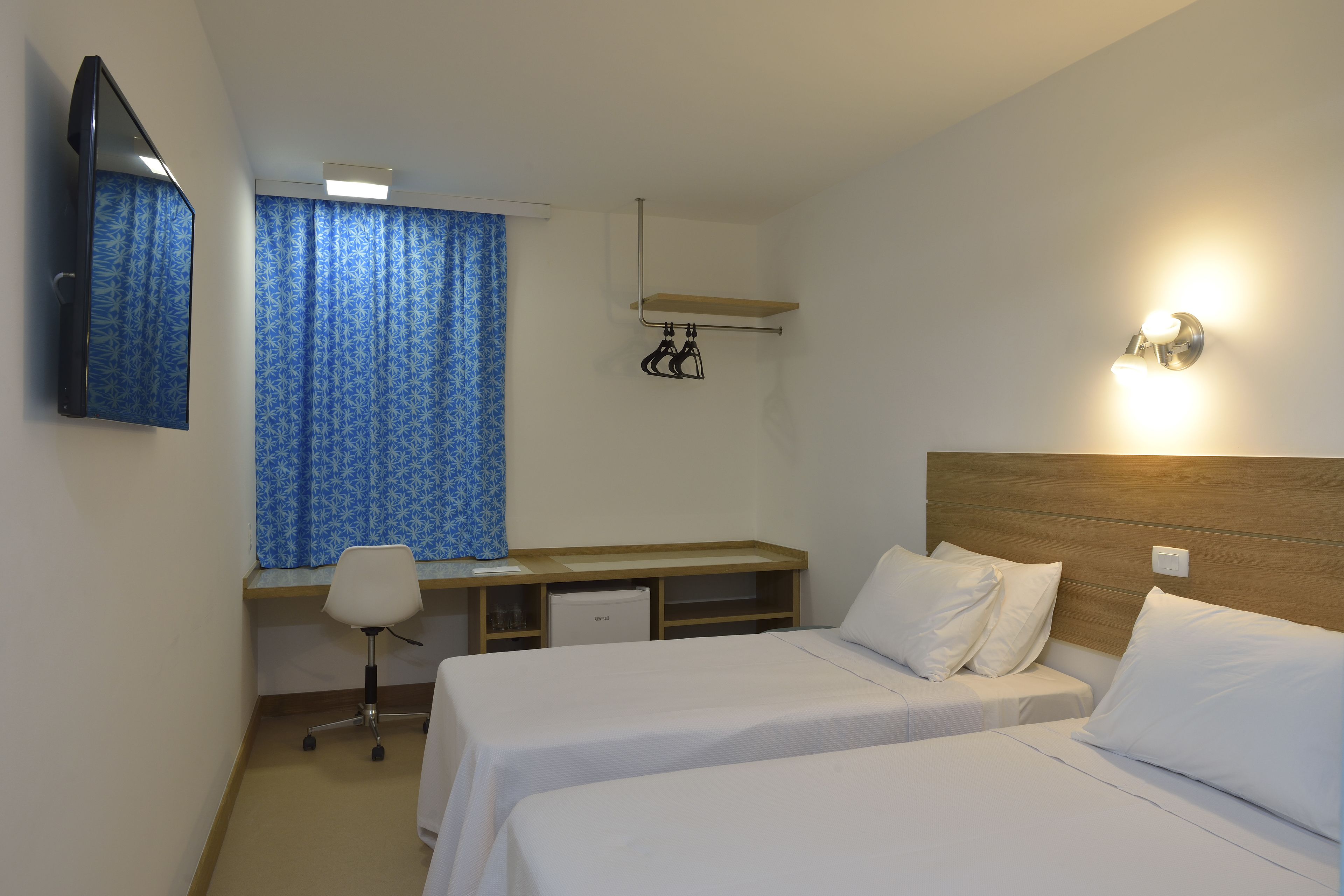 Une chambre d’hôtel avec deux lits simples, un bureau avec une chaise, un petit réfrigérateur, un téléviseur fixé au mur et une fenêtre avec des rideaux bleus.
