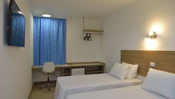 Quarto de hotel com duas camas de solteiro, uma escrivaninha com cadeira, um pequeno frigorífico, uma televisão presa à parede e uma janela com cortinas azuis.