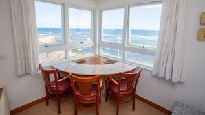 A room with a round table and four chairs, large windows with ocean views, white curtains, and a ceiling light. - Rede Andrade Express (Salvador)