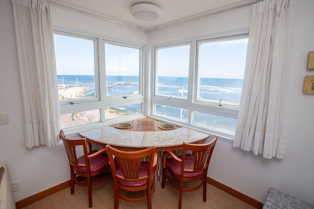 Una habitación con una mesa redonda y cuatro sillas, ventanas amplias con vistas al océano, cortinas blancas y una lámpara en el techo.