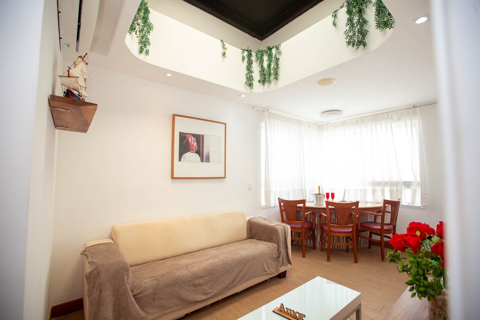 A living room with a sofa, a dining table with chairs, a framed picture on the wall, and a window with curtains.