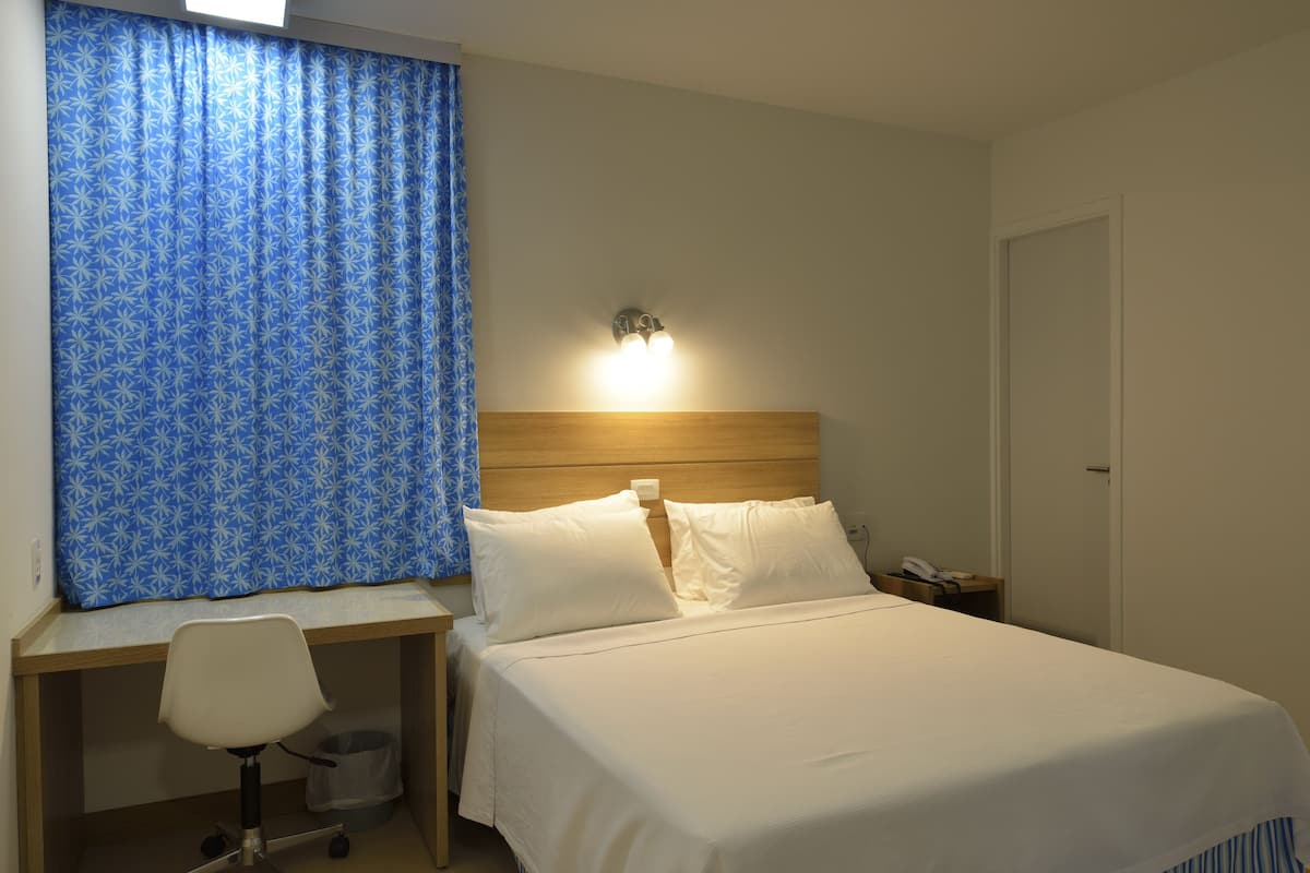 Habitación de hotel con cama, escritorio con silla, mesita de noche y una lámpara de pared.