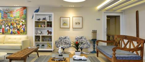 A living room with a sofa, coffee table, and chairs, a bookshelf with decorative items, and a painting on the wall.