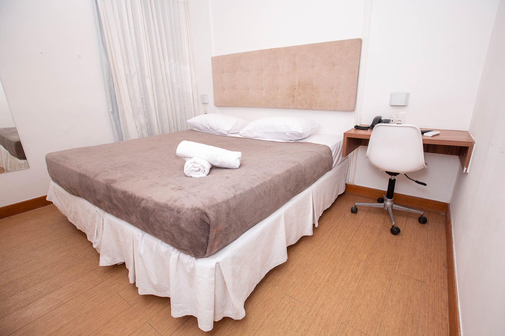 Uma cama bem arrumada com um cobertor marrom e toalhas dobradas, uma escrivaninha branca com uma cadeira, e um chão de madeira.