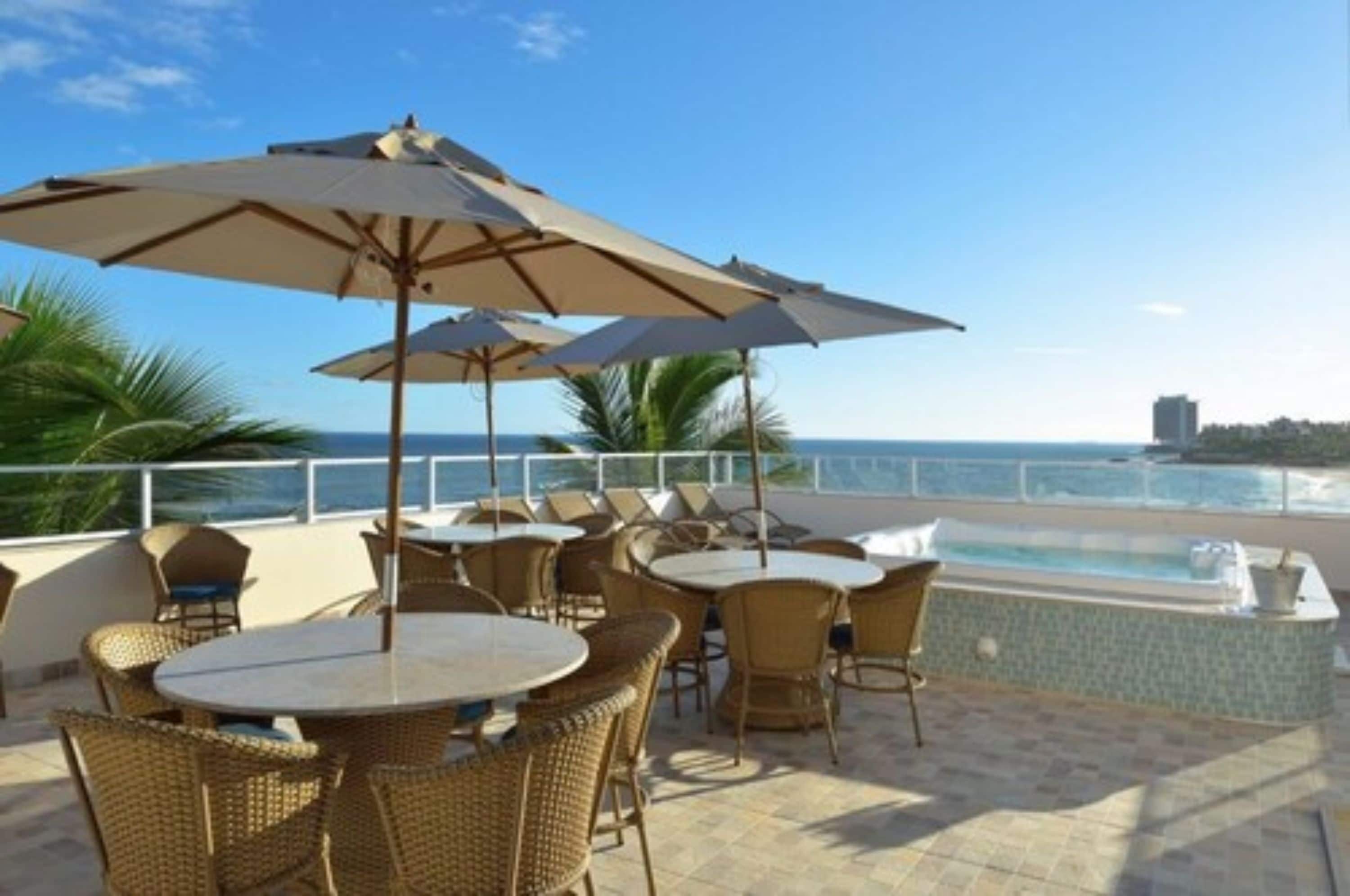 Terraço no terraço com mesas redondas e cadeiras de vime, uma piscina e vista para o oceano.