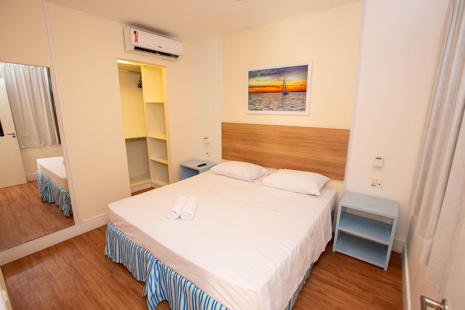 Een hotelkamer met een bed, nachtkastje, garderobe, airconditioning en een schilderij van een zonsondergang boven het water.
