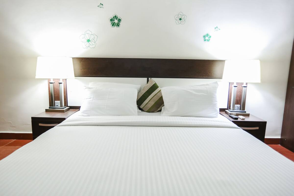 A neatly made bed with white linens and a striped pillow, flanked by two bedside tables with lamps, in a room with a simple wooden headboard and floral wall decals.